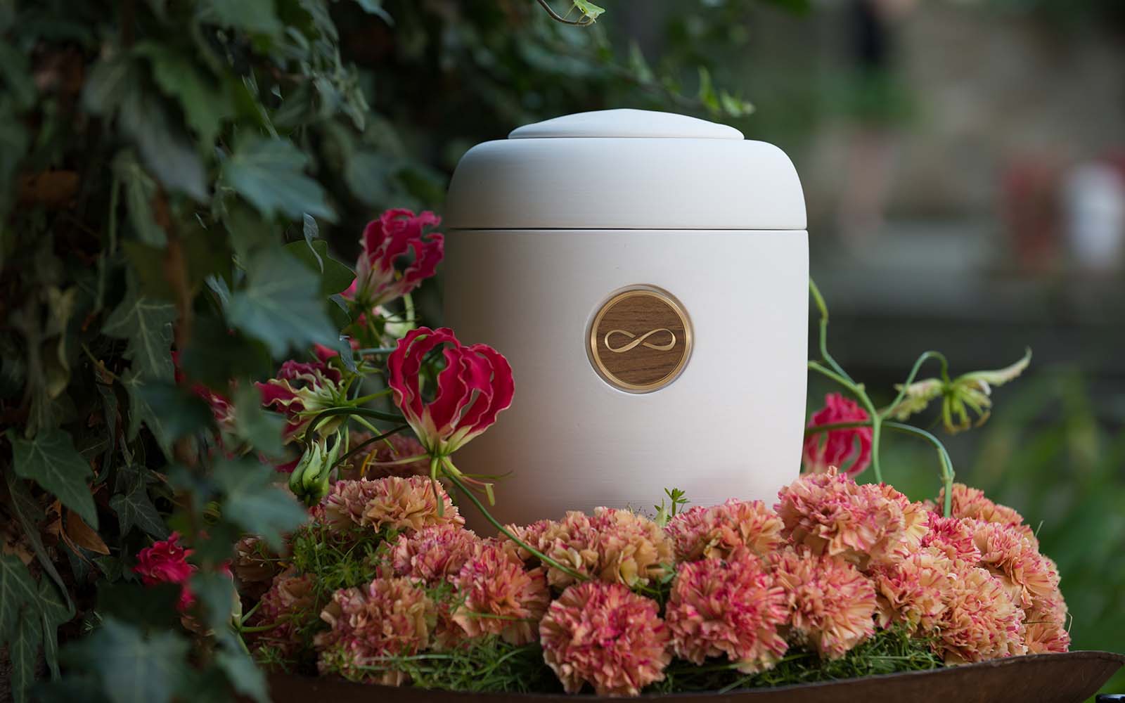 Cremefarbene Urne mit glänzendem Messing-Holz-Medaillon, eingravierte Unendlichkeits-8, umgeben von rosa Blumenkranz.