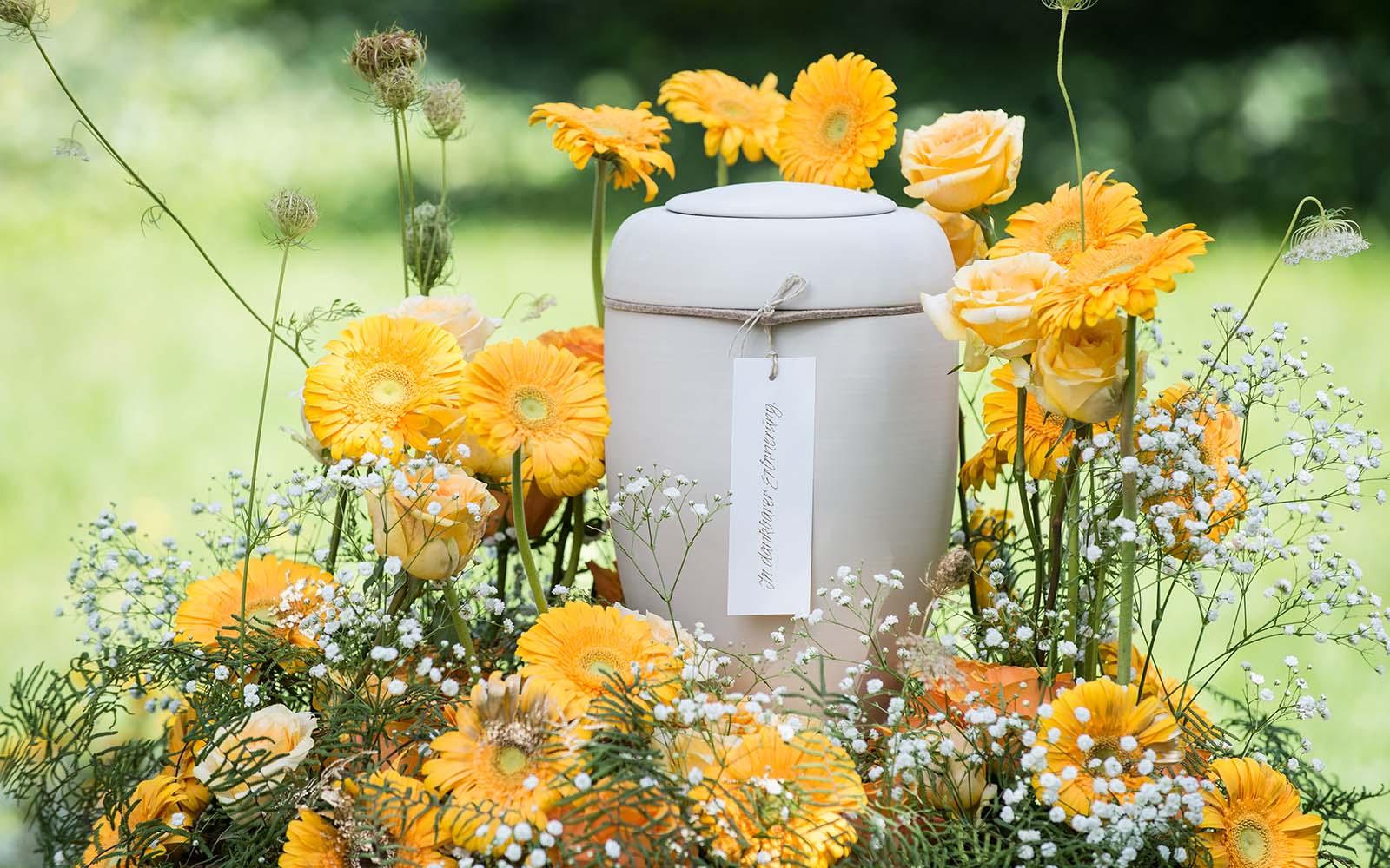 Eine sandfarbene, biologisch abbaubare Urne mit Papieranhänger steht umgeben von gelben Blumen.