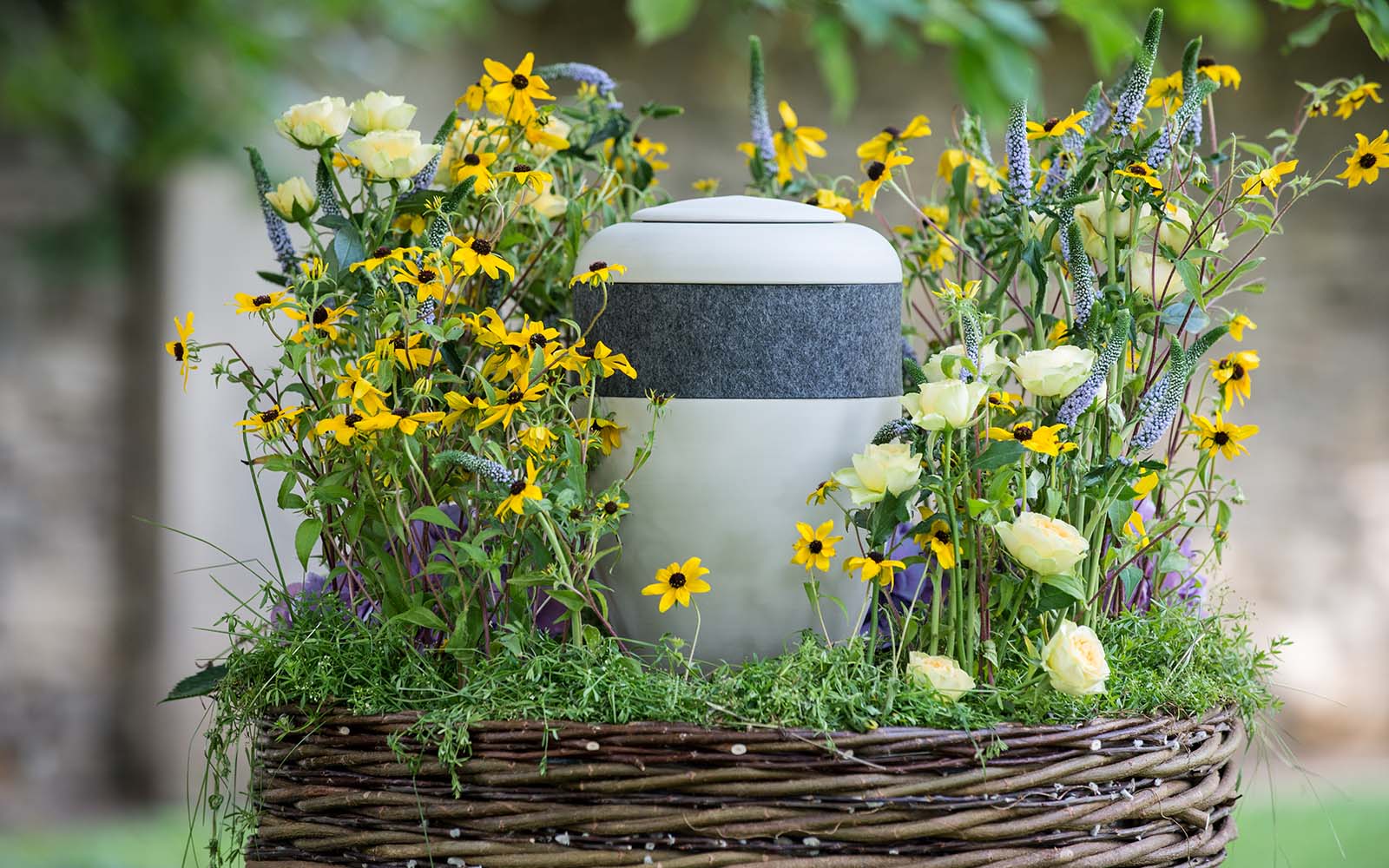 Eine schlichte, weiße Bio-Urne mit dunkelgrauem Filzring, umgeben von farbenfrohen Blumen auf einem Weidenkranz.