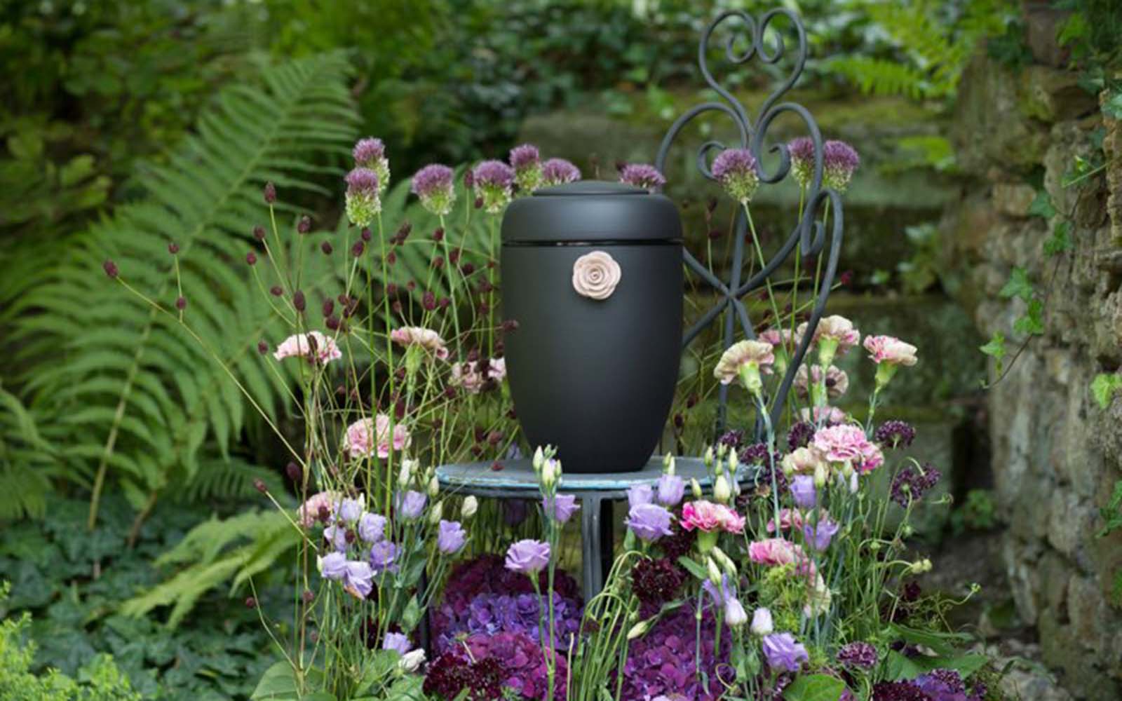 Schwarze Design-Urne mit glänzendem Ring und Tonrose, umgeben von Blumen.