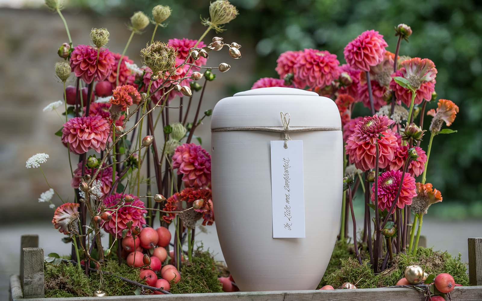 Biologisch abbaubare Urne auf Moos, umgeben von Blumen, trägt die Inschrift 'In Liebe und Dankbarkeit'