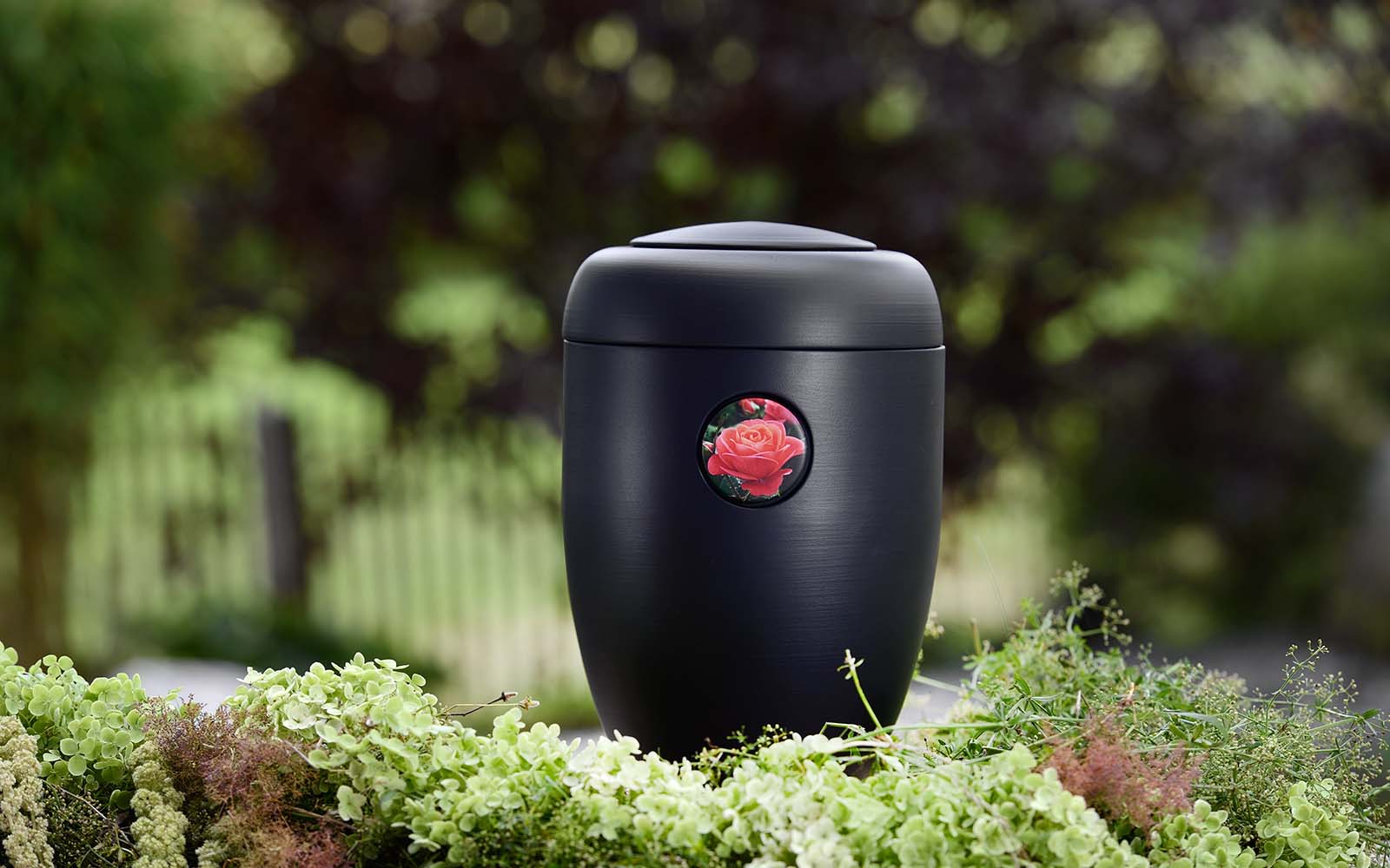 Schwarze, matte Design-Urne mit lachsfarbener Rose-Button, umrahmt von grünen Blumen.